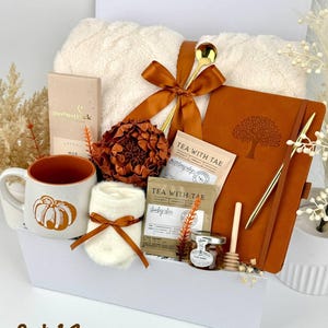 May include: A gift set containing a cream-coloured fuzzy blanket tied with a rust-coloured ribbon, a ceramic mug with a pumpkin graphic, a rust-coloured notebook with a tree emblem, a gold pen, tea sachets, a small honey jar, and a wooden dipper.