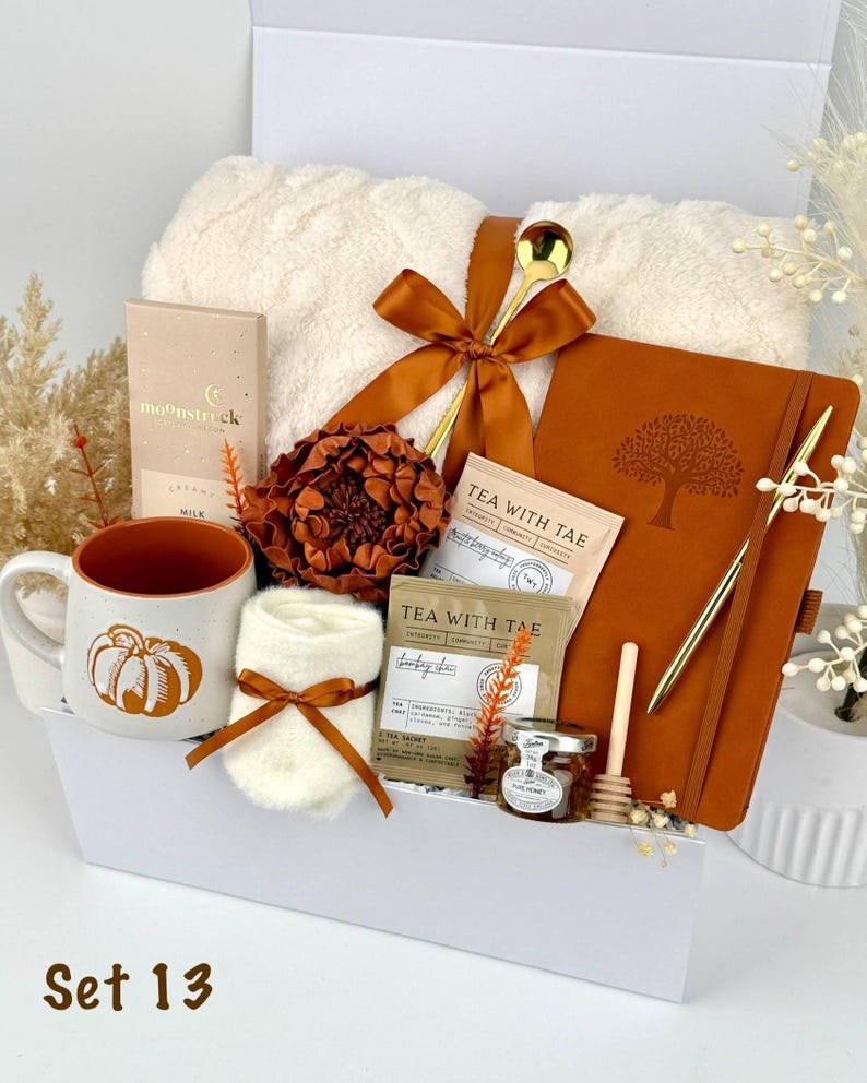 May include: A gift set containing a cream-coloured fuzzy blanket tied with a rust-coloured ribbon, a ceramic mug with a pumpkin graphic, a rust-coloured notebook with a tree emblem, a gold pen, tea sachets, a small honey jar, and a wooden dipper.