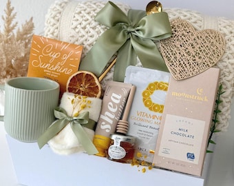 May include: A white gift box containing a cream-colored knit blanket, a sage green ribbed mug, a Cup of Sunshine tea box, a shea butter tube, a honey jar with a dipper, a Vitamin C sheet mask, a milk chocolate bar, and a decorative heart.