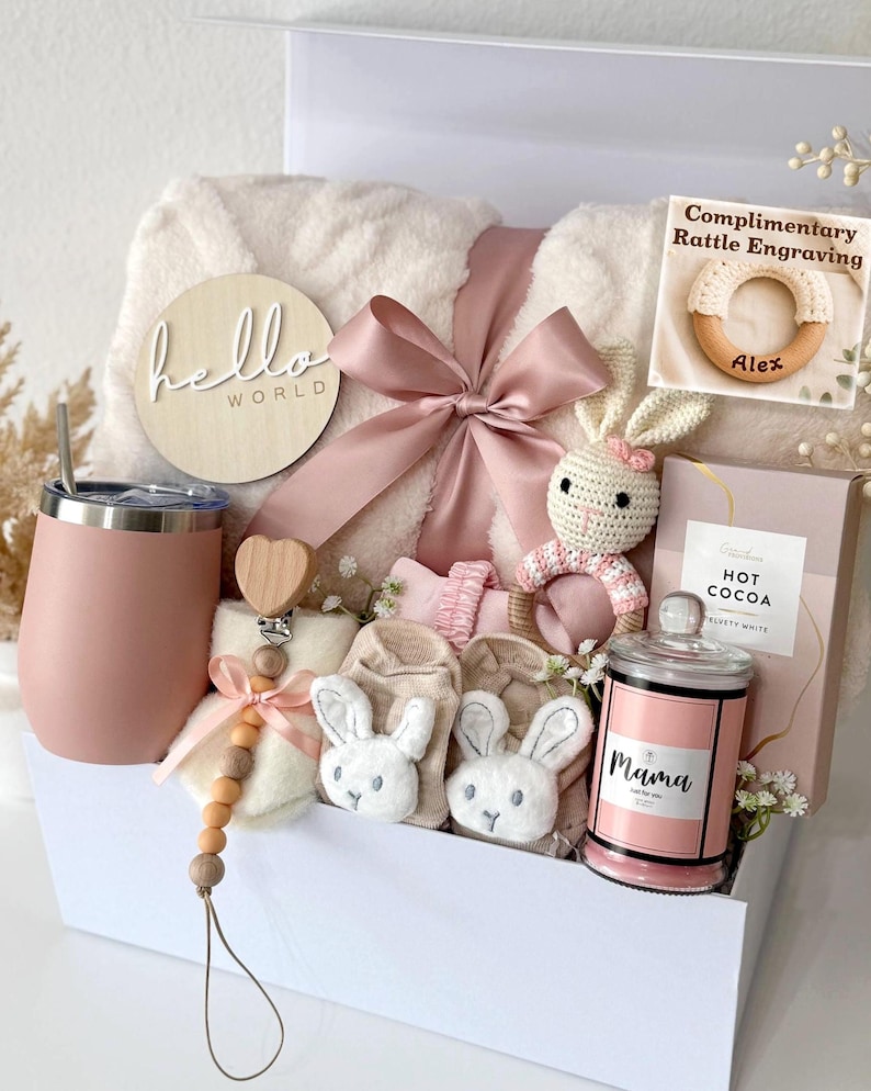 May include: A gift box containing a cream-colored plush blanket with a pink ribbon, a pink tumbler, a wooden 'hello world' sign, a crochet bunny rattle, a pacifier clip with wooden beads, baby socks with bunny faces, and a candle labeled 'Mama'.