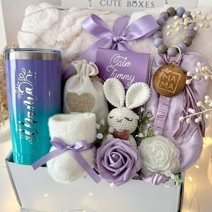 May include: A gift box containing a purple and teal ombre tumbler with the word Mama, a white fuzzy blanket, a purple sleep mask with a beaded keychain, a small crochet bunny, a burlap pouch, a white candle, and a purple rose flower.