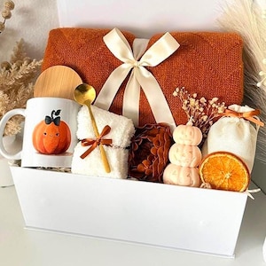May include: A white rectangular gift basket containing a folded burnt orange knit blanket with a cream ribbon, a white mug with a pumpkin illustration, a gold spoon, a white fuzzy cloth, a stacked pumpkin candle, a dried orange slice, and a pouch.