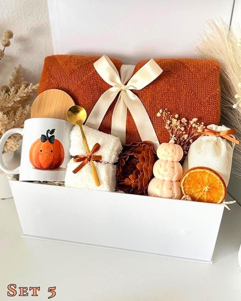 May include: A white rectangular gift basket containing a folded burnt orange knit blanket with a cream ribbon, a white mug with a pumpkin illustration, a gold spoon, a white fuzzy cloth, a stacked pumpkin candle, a dried orange slice, and a pouch.