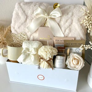 Puede incluir: Una caja de regalo blanca llena de art&iacute;culos de cuidado personal. Incluye una manta suave, una taza, una vela, sales de ba&ntilde;o, t&eacute; y una rosa decorativa. La caja est&aacute; atada con un lazo de sat&eacute;n color crema. El texto "Set 10" est&aacute; en la parte inferior.