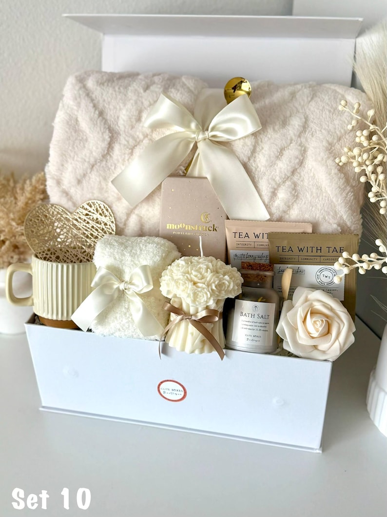 Puede incluir: Una caja de regalo blanca llena de art&iacute;culos de cuidado personal. Incluye una manta suave, una taza, una vela, sales de ba&ntilde;o, t&eacute; y una rosa decorativa. La caja est&aacute; atada con un lazo de sat&eacute;n color crema. El texto "Set 10" est&aacute; en la parte inferior.