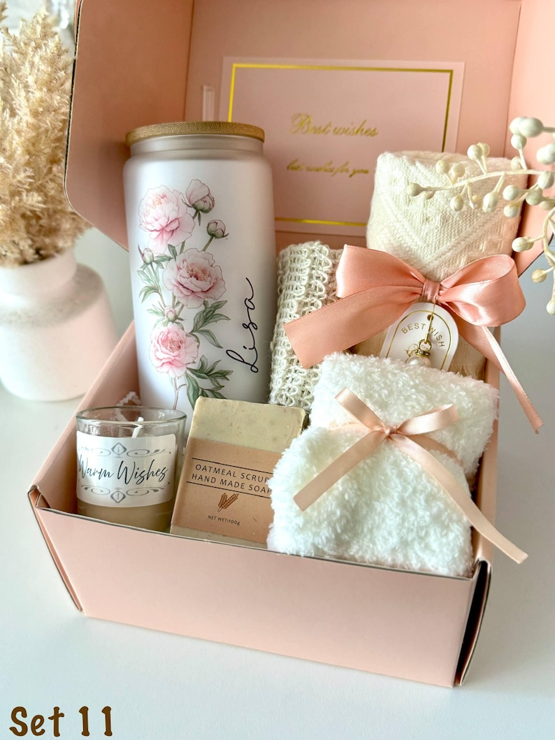May include: A gift box containing a frosted glass tumbler with a pink floral design and the name Lisa, a candle labeled Warm Wishes, a bar of oatmeal scrub handmade soap, a textured white cloth, and a plush white item tied with a peach ribbon.