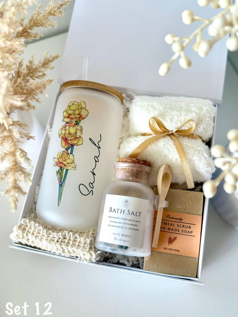 May include: A white gift box contains a frosted glass tumbler with a floral design and the name "Sarah", a jar of bath salt, a bar of oatmeal soap, two white washcloths tied with a gold ribbon, and a small wooden spoon. The text "Set 12" is at the bottom.