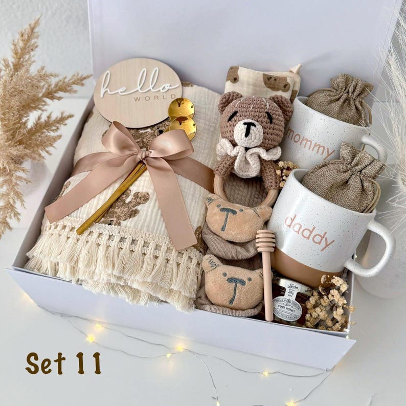 K&ouml;nnte beinhalten: Eine Baby-Geschenkbox mit einer Decke, einem Holzschild mit der Aufschrift "hello world", einem H&auml;kelb&auml;ren, zwei Tassen mit den Aufschriften "mommy" und "daddy", Babysocken und Honig. Die Box ist wei&szlig; und tr&auml;gt den Text "Set 11".