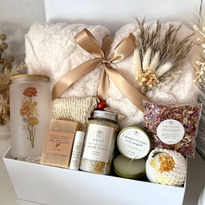 May include: A white gift box filled with self-care items. Includes a fluffy cream-colored towel tied with a gold ribbon, a frosted glass jar with floral design, soap, bath salts, a candle, and a bath bomb. The text "Set 2" is visible.