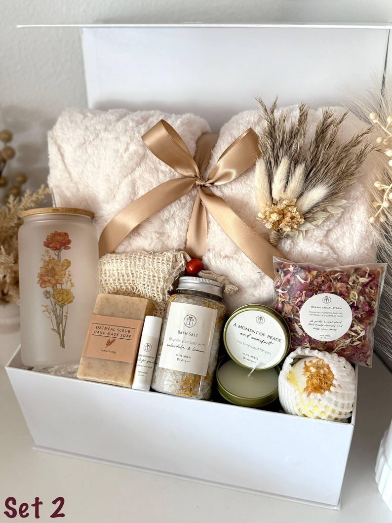 May include: A white gift box filled with self-care items. Includes a fluffy cream-colored towel tied with a gold ribbon, a frosted glass jar with floral design, soap, bath salts, a candle, and a bath bomb. The text "Set 2" is visible.