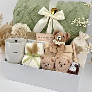 May include: A gift set containing a sage green textured blanket with a cream ribbon, a speckled ceramic mug labeled mom, two tea packets, a crochet bear rattle labeled Noah, two small plush bear faces, a honey jar with a dipper, and a fabric pouch.