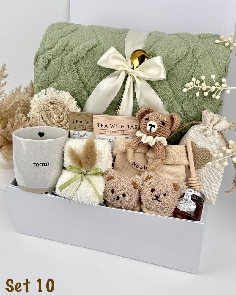 May include: A gift set containing a sage green textured blanket with a cream ribbon, a speckled ceramic mug labeled mom, two tea packets, a crochet bear rattle labeled Noah, two small plush bear faces, a honey jar with a dipper, and a fabric pouch.