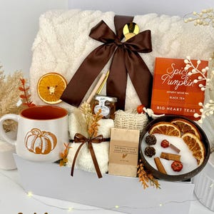 May include: A gift set containing a cream-colored textured throw blanket tied with a brown ribbon, a white mug with an orange pumpkin graphic, a box of Spicy Pumpkin black tea, a small jar of honey, oatmeal soap, and a candle in a coconut shell.