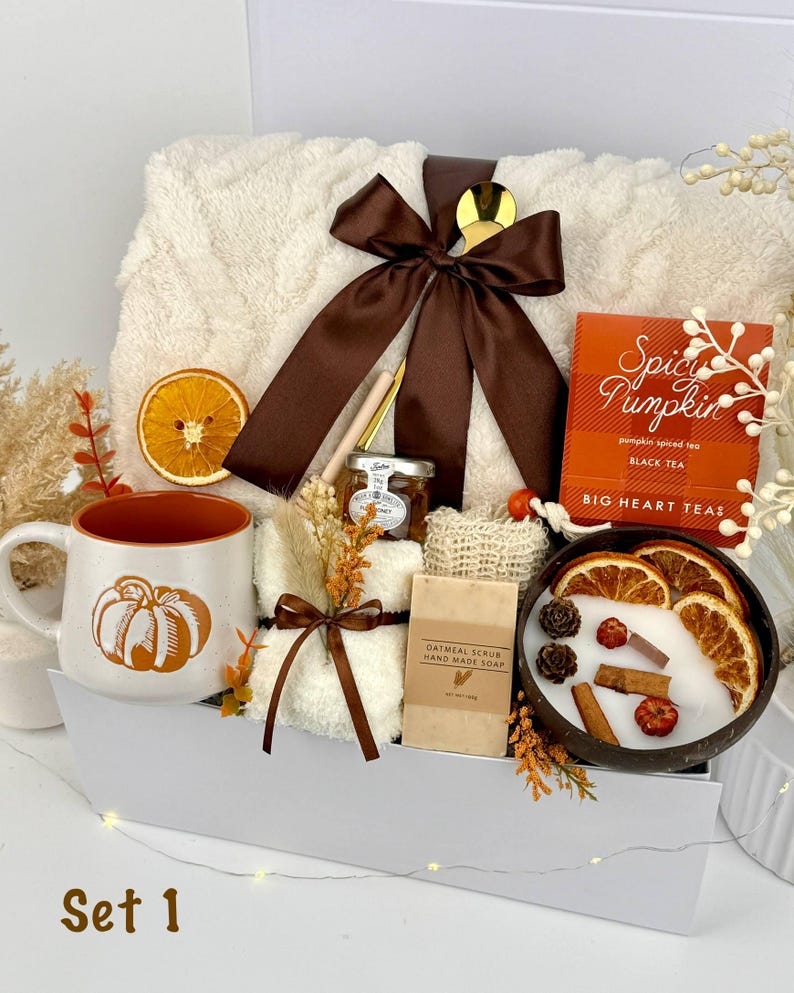 May include: A gift set containing a cream-colored textured throw blanket tied with a brown ribbon, a white mug with an orange pumpkin graphic, a box of Spicy Pumpkin black tea, a small jar of honey, oatmeal soap, and a candle in a coconut shell.