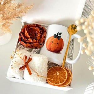 May include: A white gift box containing a white mug with a cartoon pumpkin illustration, a rust-colored felt flower, a pair of white fuzzy socks tied with a rust ribbon, a gold-toned spoon, and two dried orange slices. The text SET 7 is visible.
