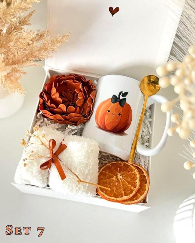 May include: A white gift box containing a white mug with a cartoon pumpkin illustration, a rust-colored felt flower, a pair of white fuzzy socks tied with a rust ribbon, a gold-toned spoon, and two dried orange slices. The text SET 7 is visible.