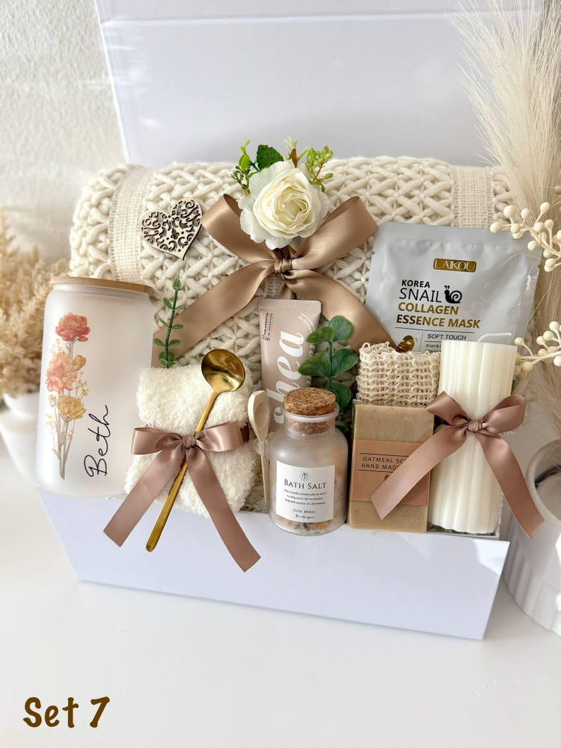 May include: A gift set in a white box, featuring a cream-colored blanket, a candle with floral design, bath salts, a face mask, soap, and a small towel. The set is decorated with ribbons and a wooden heart. The text "Set 7" is visible.