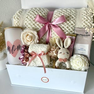 K&ouml;nnte beinhalten: Eine wei&szlig;e Geschenkbox gef&uuml;llt mit einer cremefarbenen Strickdecke, die mit einem rosa Band gebunden ist. Die Box enth&auml;lt auch eine Kerze mit dem Wort "Mom", eine wei&szlig;e Rose, ein kleines Handtuch, ein H&auml;schen und andere Artikel.