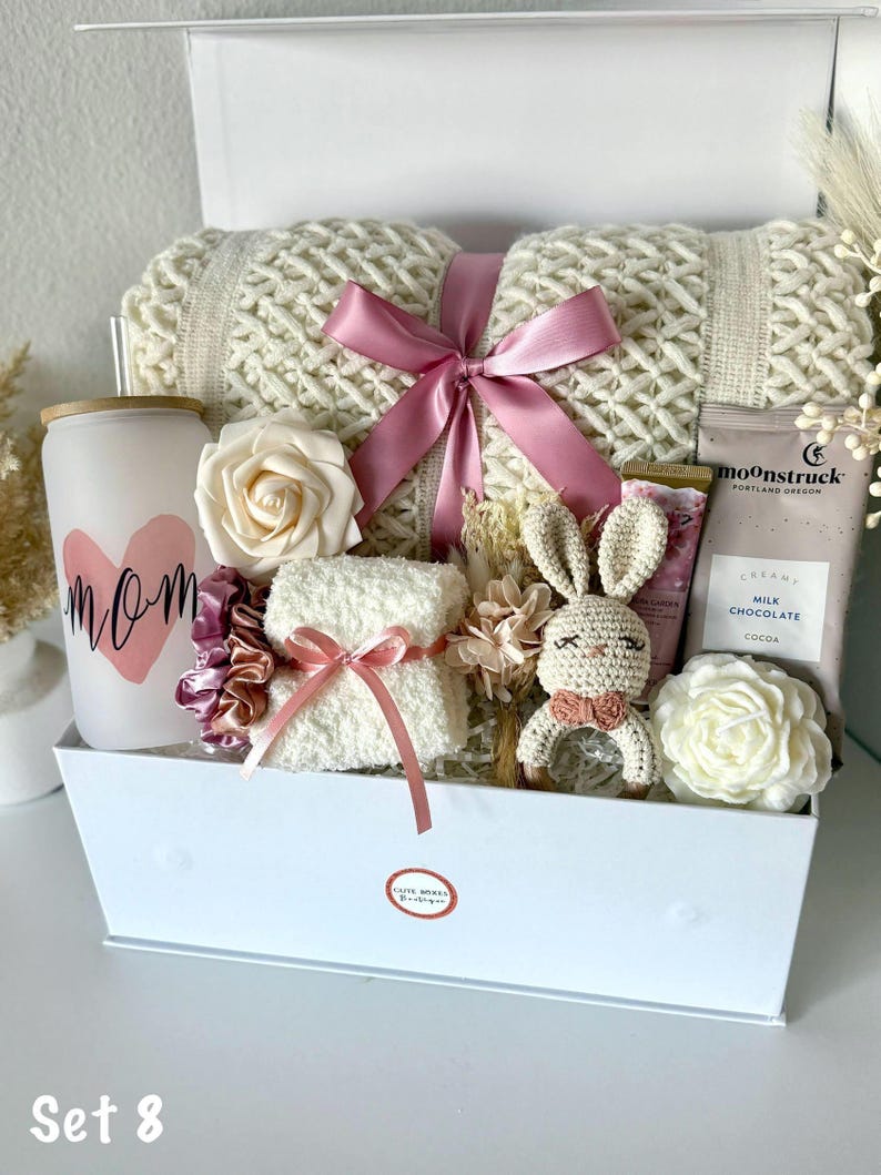 K&ouml;nnte beinhalten: Eine wei&szlig;e Geschenkbox gef&uuml;llt mit einer cremefarbenen Strickdecke, die mit einem rosa Band gebunden ist. Die Box enth&auml;lt auch eine Kerze mit dem Wort "Mom", eine wei&szlig;e Rose, ein kleines Handtuch, ein H&auml;schen und andere Artikel.