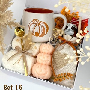 May include: A gift box containing a speckled ceramic mug with an orange pumpkin graphic, a gold spoon, a stack of white fuzzy socks, a stack of three pale orange pumpkin-shaped candles, a small jar of honey, and a burlap bag with a heart patch.