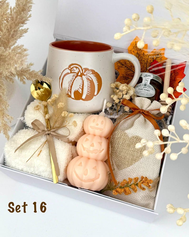 May include: A gift box containing a speckled ceramic mug with an orange pumpkin graphic, a gold spoon, a stack of white fuzzy socks, a stack of three pale orange pumpkin-shaped candles, a small jar of honey, and a burlap bag with a heart patch.