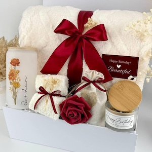 May include: A gift set containing a white fuzzy blanket tied with a dark red ribbon, a frosted glass tumbler with floral print and the name Jordan, a small white towel, a beige drawstring pouch, a dark red rose, and a candle with a bamboo lid.