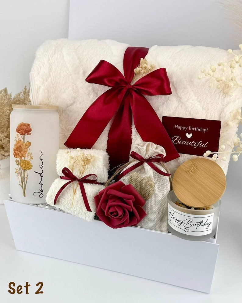 May include: A gift set containing a white fuzzy blanket tied with a dark red ribbon, a frosted glass tumbler with floral print and the name Jordan, a small white towel, a beige drawstring pouch, a dark red rose, and a candle with a bamboo lid.
