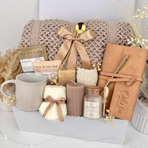 May include: A gift set containing a beige knit blanket with a gold spoon and ribbon, a ribbed mug, two Tea With Tae packets, a small soap, a textured sponge, a ribbed candle, bath salt, and a tan notebook with the name Olivia and a gold pen.