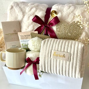 May include: A gift basket filled with self-care items. Includes a cream-colored plush blanket, a mug, chocolate, tea, a cosmetic bag with "Sarah" embroidered on it, and decorative accents. The basket is tied with a burgundy ribbon and includes a gold spoon.