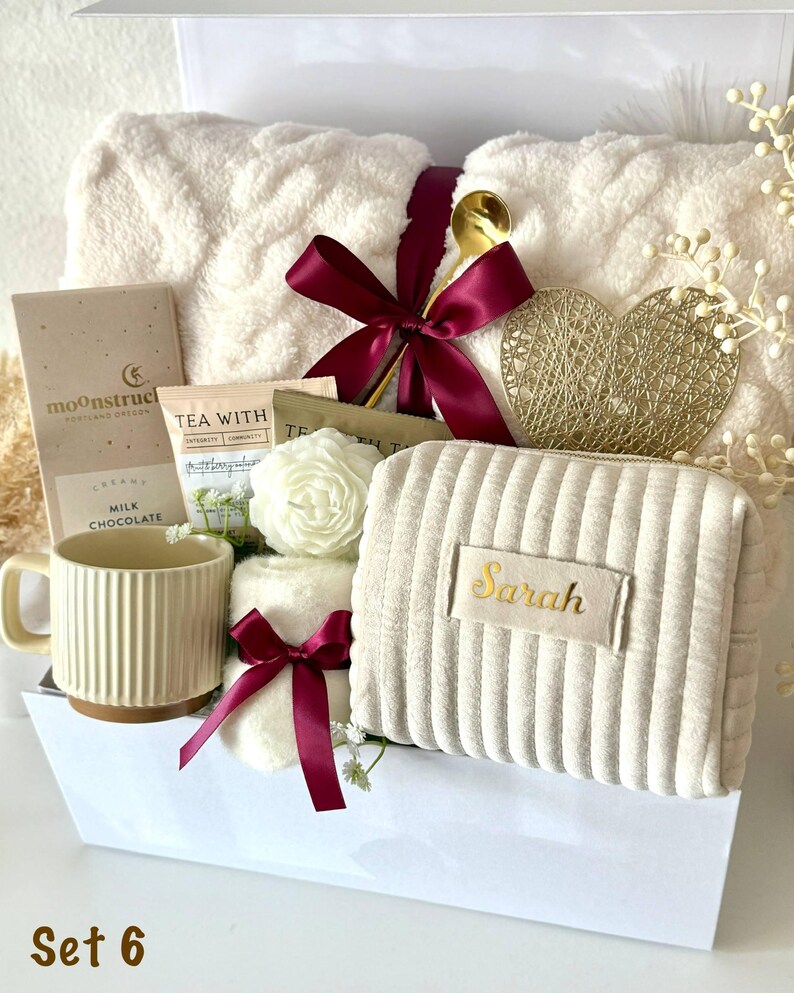 May include: A gift basket filled with self-care items. Includes a cream-colored plush blanket, a mug, chocolate, tea, a cosmetic bag with "Sarah" embroidered on it, and decorative accents. The basket is tied with a burgundy ribbon and includes a gold spoon.