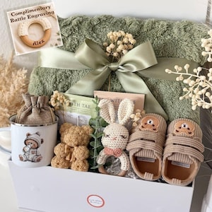 May include: A gift box containing a sage green plush blanket with a satin ribbon, a white enamel mug with a bear illustration, a small burlap pouch, a bear-shaped rose figure, a crocheted bunny rattle, and a pair of beige corduroy baby shoes.