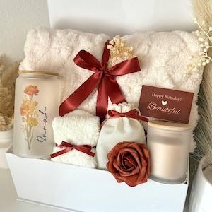 May include: A white gift box contains a fluffy cream-colored towel, a candle, a small towel, a small bag, and a decorative rose. A red ribbon and a card that says "Happy Birthday! Beautiful" are included. The candle has a floral design and the name "Sarah".