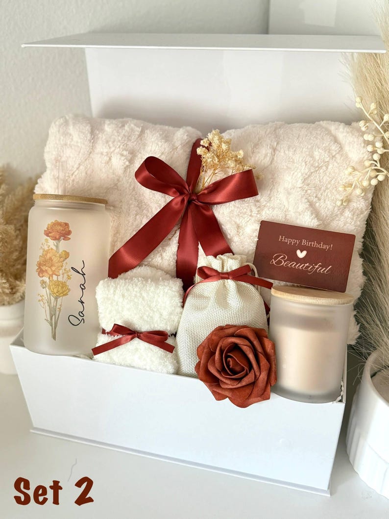 May include: A white gift box contains a fluffy cream-colored towel, a candle, a small towel, a small bag, and a decorative rose. A red ribbon and a card that says "Happy Birthday! Beautiful" are included. The candle has a floral design and the name "Sarah".
