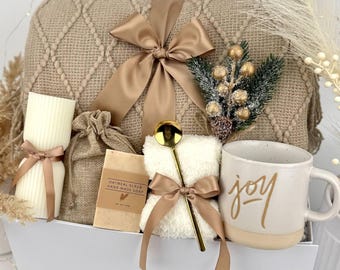 Può includere: Un set regalo contenente una coperta in maglia beige con nastro di raso, una candela a colonna a coste, un sacchetto di juta, una saponetta esfoliante all'avena, un cucchiaio dorato su un panno bianco soffice e una tazza con la scritta joy.