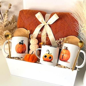 May include: A white gift box containing a folded orange knit blanket tied with a cream ribbon, three white mugs with pumpkin illustrations, three bamboo lids, two gold spoons, a small orange fabric pumpkin, and a stacked pumpkin candle.