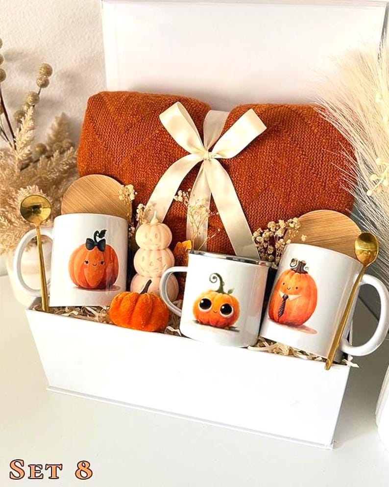 May include: A white gift box containing a folded orange knit blanket tied with a cream ribbon, three white mugs with pumpkin illustrations, three bamboo lids, two gold spoons, a small orange fabric pumpkin, and a stacked pumpkin candle.