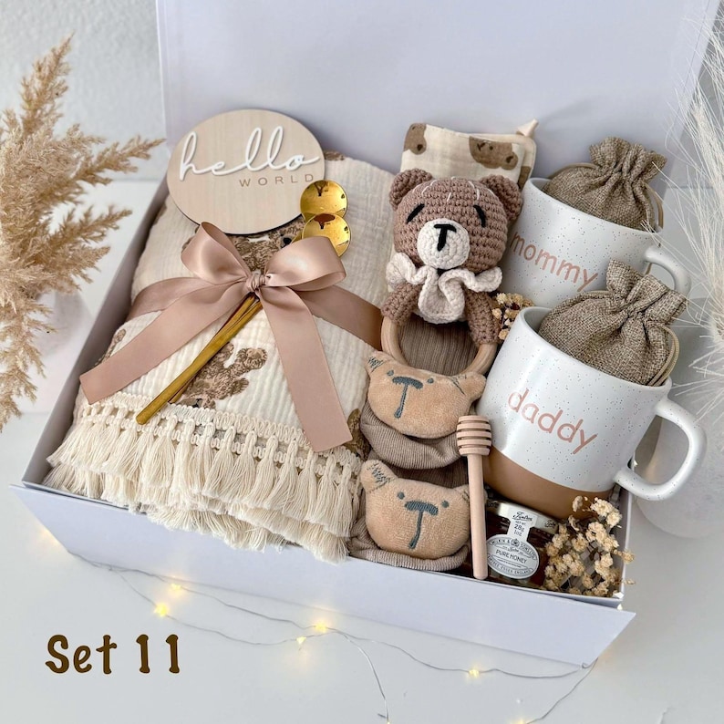 May include: A gift box containing a cream-colored fringed blanket with a brown bear pattern, a wooden hello world disc, a crocheted bear rattle, two speckled ceramic mugs labeled mommy and daddy, a honey dipper, and a small jar of honey.