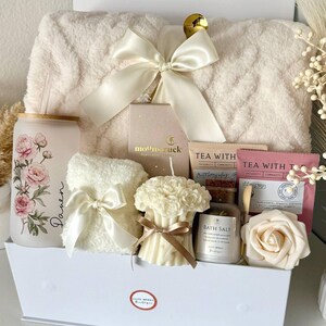 May include: A white gift box filled with self-care items. Includes a fluffy cream-colored blanket, a floral-printed glass, a small towel with a bow, a candle, bath salts, tea bags, and a decorative rose. The box is tied with a cream-colored bow.