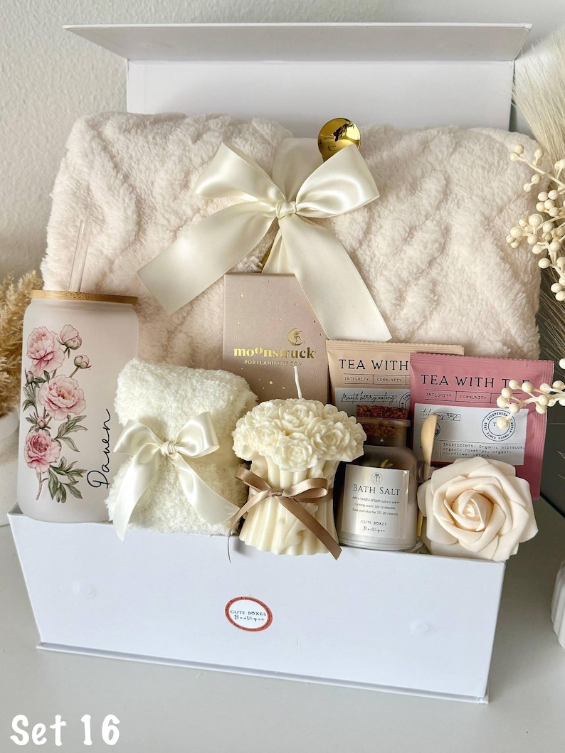 May include: A white gift box filled with self-care items. Includes a fluffy cream-colored blanket, a floral-printed glass, a small towel with a bow, a candle, bath salts, tea bags, and a decorative rose. The box is tied with a cream-colored bow.