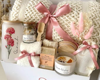 May include: A white gift box filled with self-care items. Includes a cream-colored knitted blanket, a candle, bath salts, soap, a small towel, and decorative flowers. The items are tied with pink ribbons.