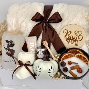 May include: A gift set containing a frosted glass tumbler with a witch graphic and the name Rashell, a cream-colored fuzzy blanket with a brown ribbon, a pair of white socks, a small jar of honey with a dipper, a ceramic pumpkin, and a candle.