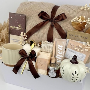 May include: A gift set containing a beige knit blanket with a brown ribbon, a ribbed cream mug, a gold spoon, a Moonstruck dark chocolate bar, Tea With Tae tea bags, a shea butter tube, oatmeal soap, a honey dipper, and a white ceramic pumpkin.