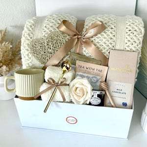Puede incluir: Una caja de regalo blanca llena de una manta de punto color crema, una cuchara dorada, una taza, bolsitas de t&eacute;, un osito de peluche, una decoraci&oacute;n en forma de coraz&oacute;n, una rosa, chocolate y miel. La caja est&aacute; atada con una cinta dorada.