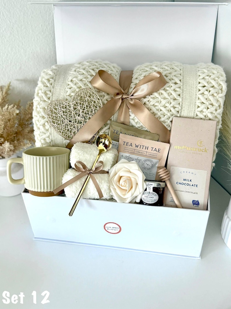 Puede incluir: Una caja de regalo blanca llena de una manta de punto color crema, una cuchara dorada, una taza, bolsitas de t&eacute;, un osito de peluche, una decoraci&oacute;n en forma de coraz&oacute;n, una rosa, chocolate y miel. La caja est&aacute; atada con una cinta dorada.