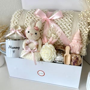 K&ouml;nnte beinhalten: Eine wei&szlig;e Geschenkbox gef&uuml;llt mit einer cremefarbenen Strickdecke, einem geh&auml;kelten Hasen, einer Tasse mit dem Text "Mommy", einer rosenf&ouml;rmigen Kerze, Badesalzen und anderen Pflegeprodukten. Die Box ist mit einem rosa Band gebunden.