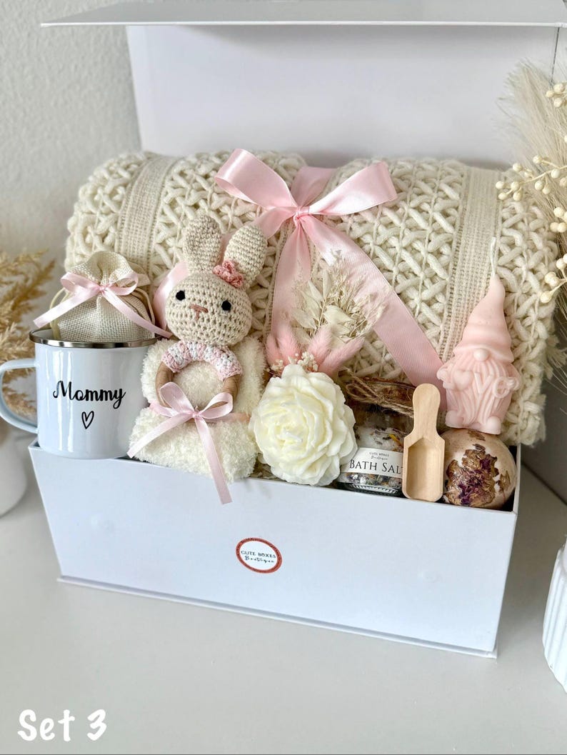 K&ouml;nnte beinhalten: Eine wei&szlig;e Geschenkbox gef&uuml;llt mit einer cremefarbenen Strickdecke, einem geh&auml;kelten Hasen, einer Tasse mit dem Text "Mommy", einer rosenf&ouml;rmigen Kerze, Badesalzen und anderen Pflegeprodukten. Die Box ist mit einem rosa Band gebunden.