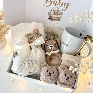 K&ouml;nnte beinhalten: Ein Baby-Geschenkset in einer wei&szlig;en Box mit einer Decke mit Teddyb&auml;ren-Stickerei, einem H&auml;kelrassel, einer Tasse mit "Mama"-Text und Teddyb&auml;ren-Socken. Der Deckel der Box hat den Text "Hello Baby" und eine Teddyb&auml;ren-Illustration.