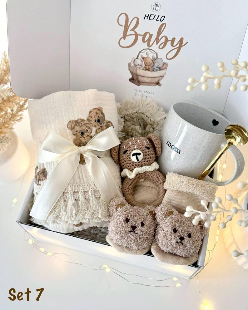 K&ouml;nnte beinhalten: Ein Baby-Geschenkset in einer wei&szlig;en Box mit einer Decke mit Teddyb&auml;ren-Stickerei, einem H&auml;kelrassel, einer Tasse mit "Mama"-Text und Teddyb&auml;ren-Socken. Der Deckel der Box hat den Text "Hello Baby" und eine Teddyb&auml;ren-Illustration.