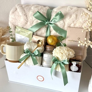 Puede incluir: Una cesta de regalo llena de art&iacute;culos de cuidado personal. Incluye una manta de felpa color crema atada con una cinta verde salvia, una taza, t&eacute;, sales de ba&ntilde;o, una vela, miel y una cuchara dorada. La cesta es blanca y est&aacute; etiquetada como "Set 7".