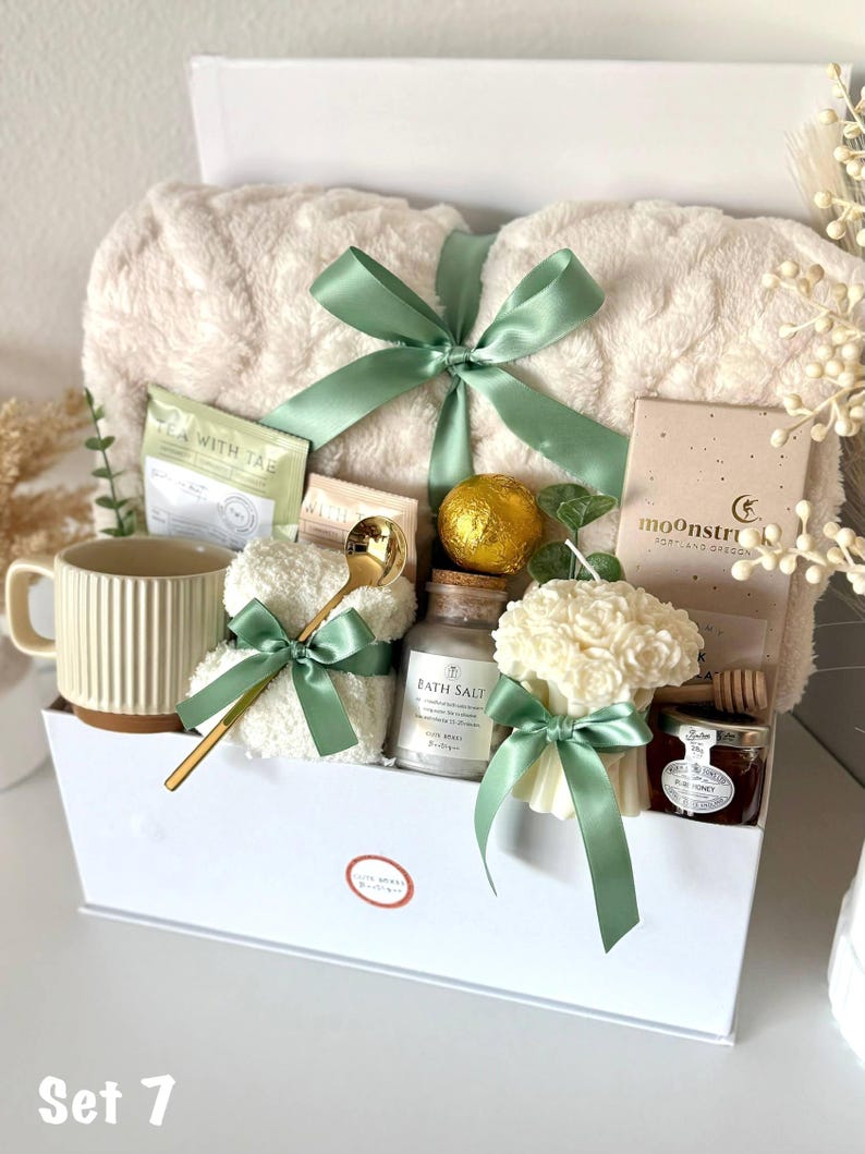 Puede incluir: Una cesta de regalo llena de art&iacute;culos de cuidado personal. Incluye una manta de felpa color crema atada con una cinta verde salvia, una taza, t&eacute;, sales de ba&ntilde;o, una vela, miel y una cuchara dorada. La cesta es blanca y est&aacute; etiquetada como "Set 7".